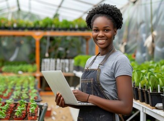 A happy young Black woman in a greenhouse, working on a laptop surrounded by lush green plants, exemplifying modern agriculture and technology integration.
