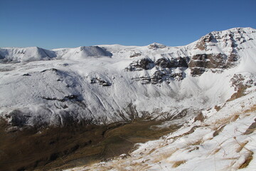 Snow blanketed mountain range creates a picturesque winter wonderland. Deep valley adds to dramatic scenery. Clear blue sky provides stunning backdrop to majestic mountain view.