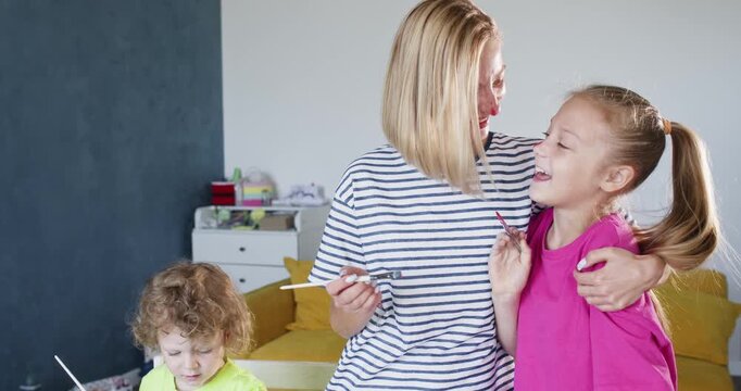 Mother applies bright red paint to tip of nose of daughter. Blonde mom and daughter spend time together. Eyes of daughter sparkle with happiness