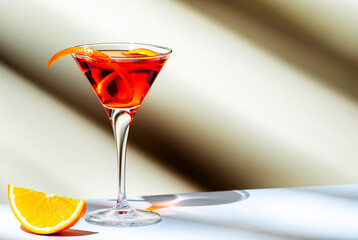 Orange cocktail with red vermouth, liqueur, bitter, citrus zest and ice in martini glass on beige background, hard light