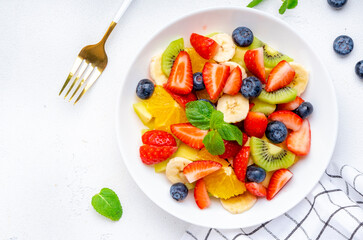 Summer vegan fruity salad with strawberries, blueberries, banana, kiwi, orange and mint on white background, top view