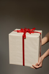 Hands delicately holding a beautifully wrapped gift box with a striking red ribbon on a neutral grey background. Holiday, celebration, gift, and special occasion concept