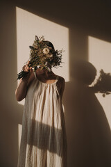 Young pretty woman in linen dress holding bouquet of flowers against white wall with hard shadows...