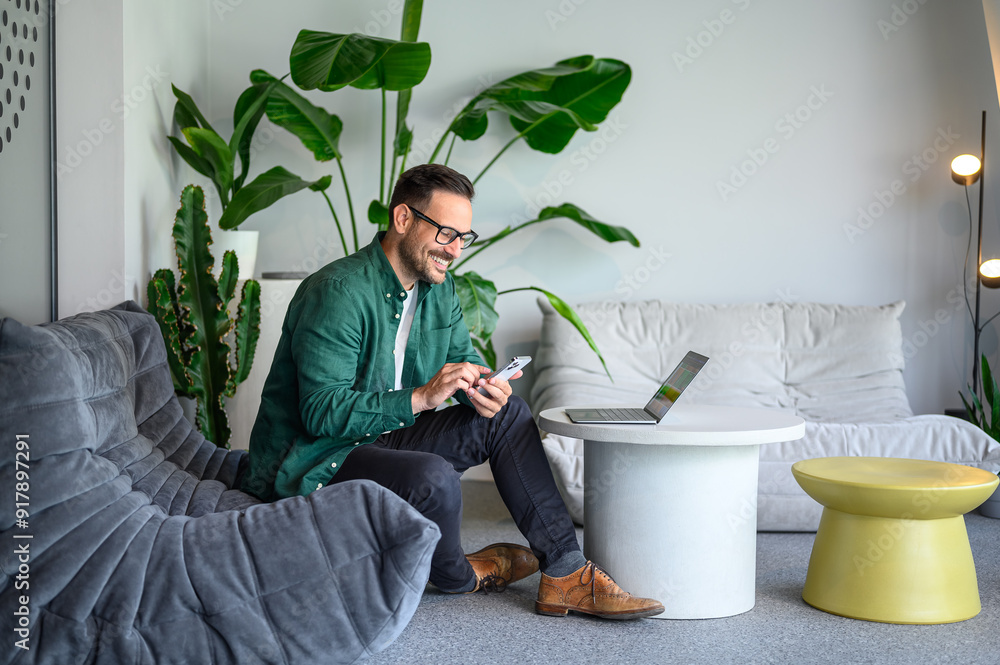 Wall mural happy manager with laptop on table using smart phone apps while sitting on sofa in modern workplace