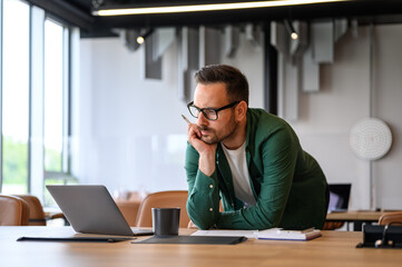 Serious young businessman with hand on chin bending over desk and reading e-mails over laptop in...