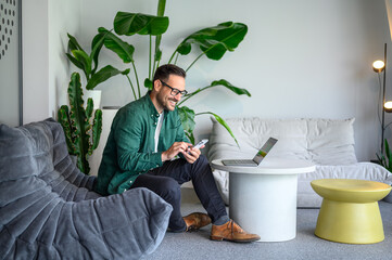 Happy manager with laptop on table using smart phone apps while sitting on sofa in modern workplace