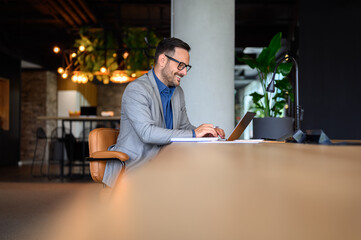 Smiling young businessman in eyeglasses working on his laptop while sitting at desk in modern office