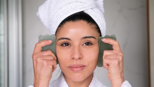 A woman is using green jade gua sha stone to massage her face after shower. She has a white towel and a white robe
