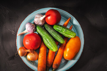 Summer vegetables on a turquoise plate on a dark gray background: tomatoes, cucumbers, carrots, garlic, onions.