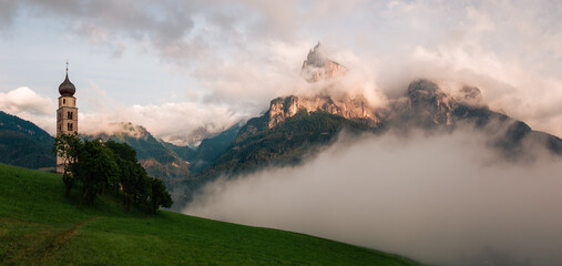 Fog envelops a serene village with a church and mountains at sunset in the Alps