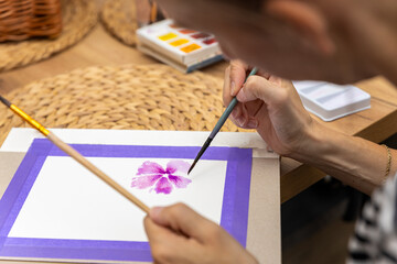 Osoba tworząca obraz akwarelowy z motywem kwiatów w odcieniach różu i fioletu. Dzieło znajduje się na podkładce z fioletową krawędzią. W tle fragment palety z farbami i brązowy, pleciony detal. © Clear Emotion