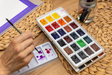 dużą paletę farb akwarelowych w kostkach, z wieloma różnorodnymi kolorami. Obok widoczna jest mniejsza, biała paletka z rozrobionymi fioletowymi i różowymi barwami, do której dłoń z pędzlem nabiera ko © Clear Emotion