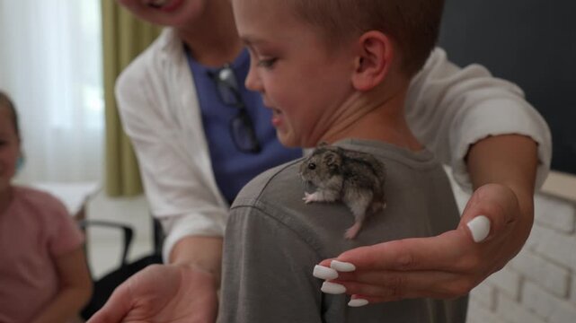 Junior school kids in classroom, woman teacher and children care for hamster in zoo corner of class, tutor and kids playing with hamster. Elementary primary education.