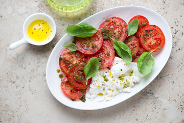 Red tomatoes served with basil oil, burrata and fresh basil on a white plate, horizontal shot on a beige granite background, high angle view