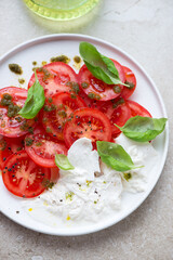 White plate with sliced red tomatoes, basil oil, fresh basil and burrata cheese, vertical shot on a beige stone surface, middle close-up