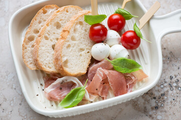 Beige serving tray with fresh ciabatta, torn prosciutto and caprese skewers, horizontal shot, middle close-up, selective focus