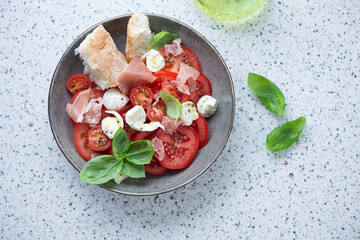 Italian caprese salad served with torn prosciutto and ciabatta, top view on a light-grey granite...