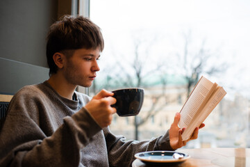 Young handsome man 20-22 year old reading book and drinking coffee in cafe. Generation Z.