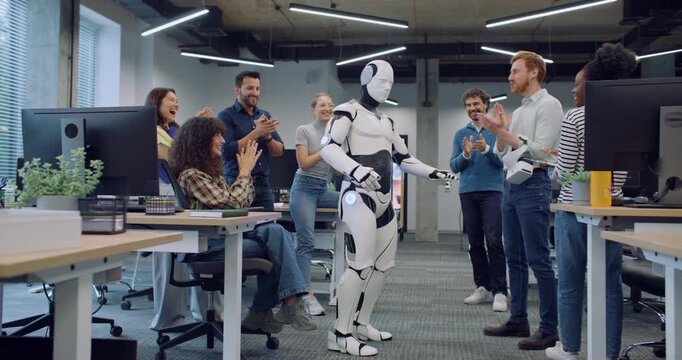 Multi-ethnic group of people surrounding their new creation in middle of large office room. Clapping together. In middle robot dancing before giving handshake to male. Showing thumbs up to camera.