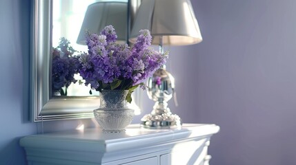 Bedroom chest by mirror; purple flowers and silver lamp on white nightstand by silver-framed mirror