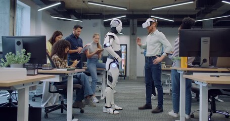 Multi-ethnic group of researchers testing new creation. Grown male wearing VR headset. Trying to operate humanoid robot with help of virtual reality glasses. Successful test. Everyone clapping.