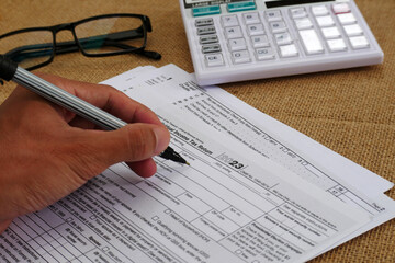 The businessman's hand is writing on a tax form, there is a calculator and glasses on the table