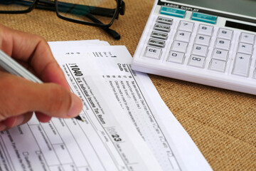 The businessman's hand is writing on a tax form, there is a calculator and glasses on the table