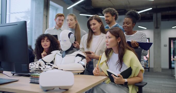 Group of multi-ethnic students surrounding robot. Humanoid sitting at desk and typing on keyboard. People using tablets and notepads. Writing down their observations. Curiosity. New mechanics.