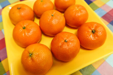 close up  top shot of nine orange citrus tangerine fruit on a yellow plate on a table with colourful table cloth