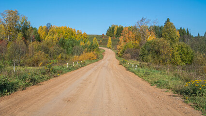 Rural dirt road in autumn. In autumn, trees with yellowed leaves grow along the country road. Trees with yellow leaves and a blue sky. Traveling by car in rural areas.