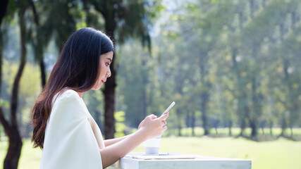 Asian businesswoman working remotely in a park using a smartphone and documents, showcasing modern...
