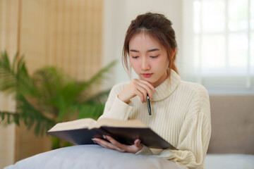 Young asian women in sweater sitting on comfortable couch and reading a book for thinking and...