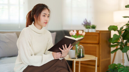 Fototapeta premium Young asian women in sweater reading a book to thinking and learning about literature story while sitting on couch to relaxation and doing activity to spending time with slow life lifestyle at home