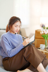 Young asian women in sweater sitting and leaning on couch to reading book and taking note for learning education while relaxation and doing activity for spending time with slow life lifestyle at home