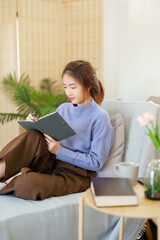 Young asian women in sweater sitting and leaning on the couch to writing notes and journey diary in notebook while relaxation and doing activity for spending time with slow life lifestyle at home