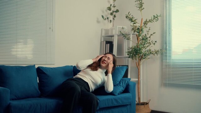 Woman in a bright living room struggling with a severe headache, expressing distress and discomfort, eventually lying down on the blue couch, clearly in pain