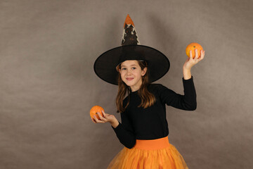 Portrait of a teenage girl in a witch hat on a gray background holding small pumpkins in her hands,...