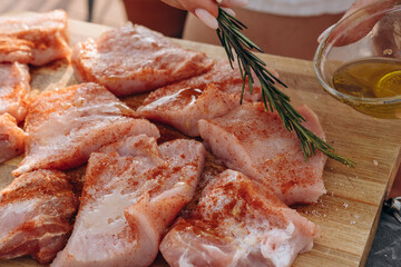 Close-up of a woman's hands adding spice marinade and olive oil  with fresh rosemary to meat steaks for cooking on a home patio grill