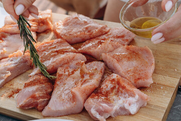 Close-up of a woman's hands adding spice marinade and olive oil  with fresh rosemary to meat steaks for cooking on a home patio grill