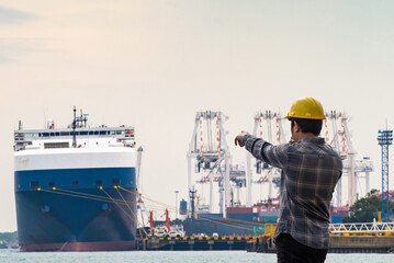 A worker in a yellow hard hat points toward a large cargo ship docked at a busy industrial port...