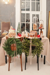 A beautiful festive table, wreaths of fir trees on the chairs. There are delicious cookies in the form of gingerbread houses and snowflakes on the table. A glass green Christmas tree and beautiful sta