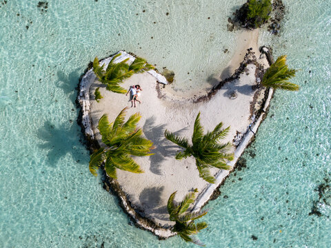 Aerial drone view of a couple laying down on a white sand beach seen on a heart shaped motu. The tropical island for the romantic moment has several palm trees and is surrounded by azure ocean water. 