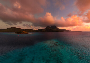Puffy pink and orange colorful clouds over Mount Otemanu on tropical island Bora Bora. Clear azure south pacific ocean water reveals a coral reef during sunrise.
