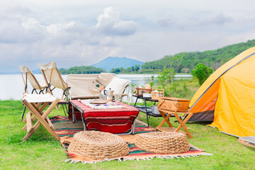 Scenic lakeside camping setup with yellow tent, colorful blankets, chairs, and picnic spread. Stunning backdrop of mountains and water. Perfect outdoor adventure scene combining comfort and nature.