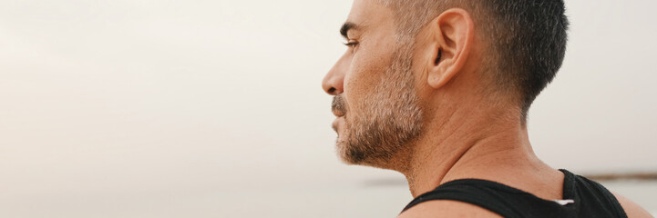 Obraz premium Close-up of middle-aged athletic man standing on the beach after workout and looking at the seascape, panorama