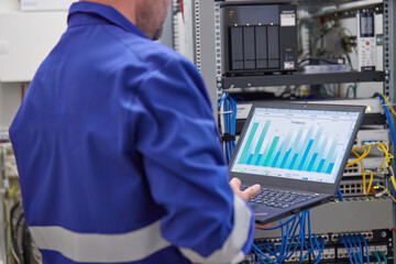 Engineer Monitoring System Performance on Laptop in High-Tech Industrial Environment – Real-Time Data Analysis for Optimized Operations and Maintenance