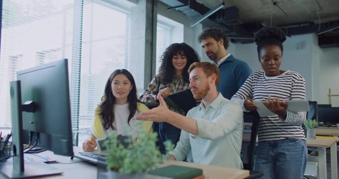 Positive multi-ethnic team working together at one desk. Listening to young man in middle describing something on screen and making gestures with his hand. Everyone looking at camera and smiling.