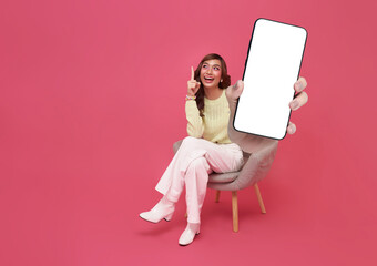 Full length beautiful Asian teen woman using smartphone and sitting on chair isolated on pink copy...