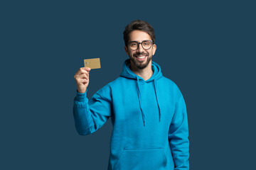 A young man wearing a bright blue hoodie stands confidently, smiling while holding up a gold card....