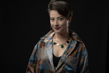 A serene woman with hoop earrings, a necklace, and short hair, wearing a multi-colored shirt, smiles gently at the camera, representing confidence and modern fashion.